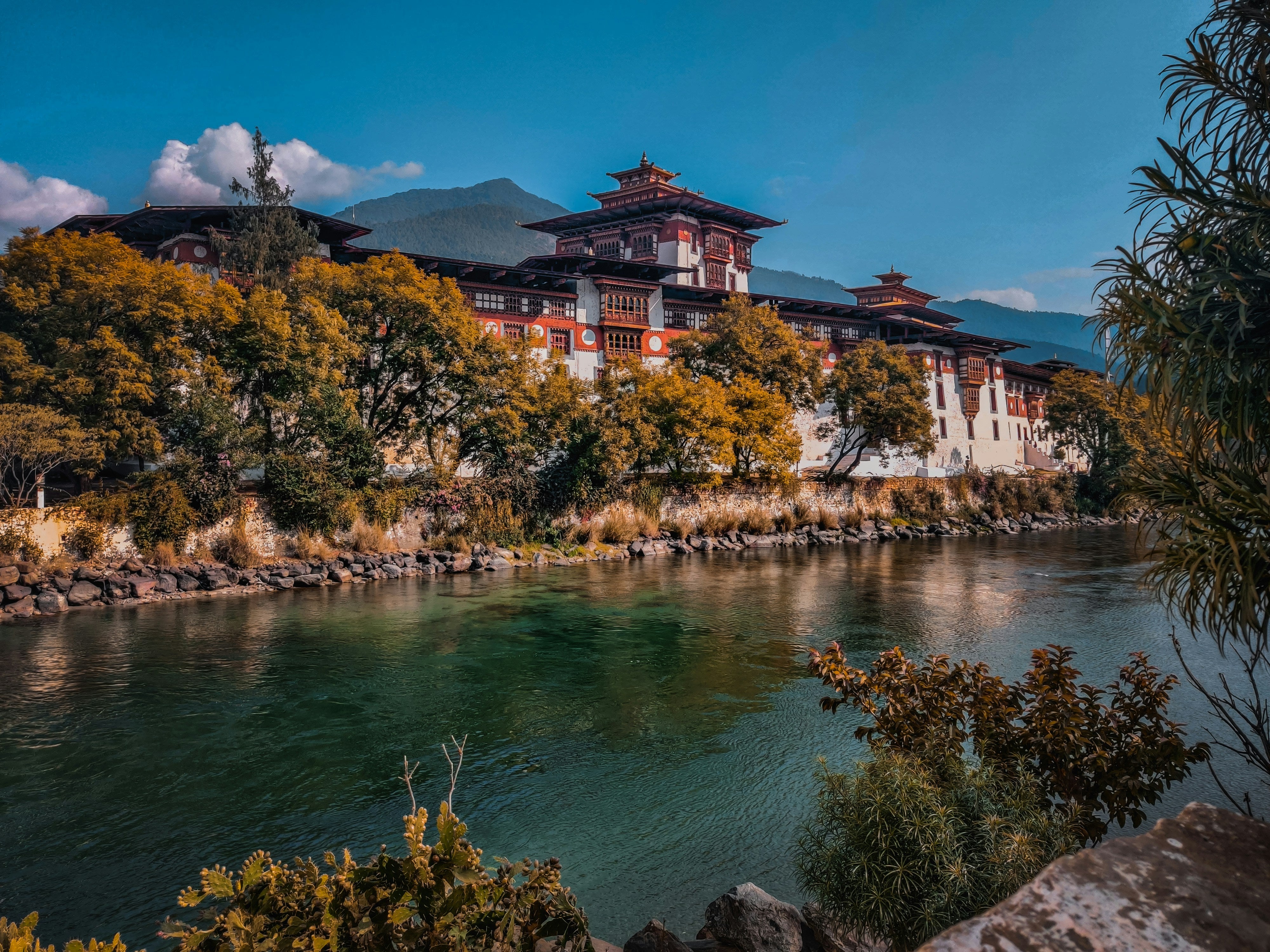 Image of A scenic landscape photograph of traditional Tibetan-style buildings with red roofs situated beside a river, surrounded by autumn-colored trees and distant mountains under a clear blue sky.